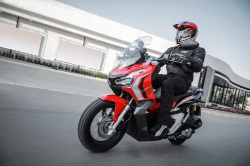 honda adv 150 vermelha sendo pilotada na cidade com imóvel comercial ao fundo